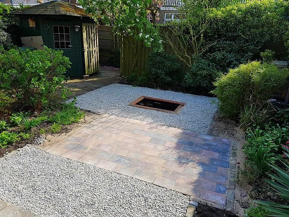 Een tuin met een stenen terras, grind en een centrale vuurkuil, omgeven door struiken en een schuur.