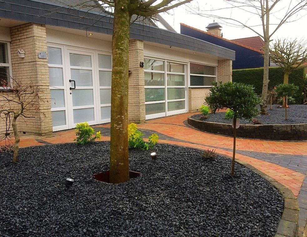 Een tuin met zwart grind, een boom voor het huis, witte garagedeuren, een bakstenen oprit en kleine struiken.