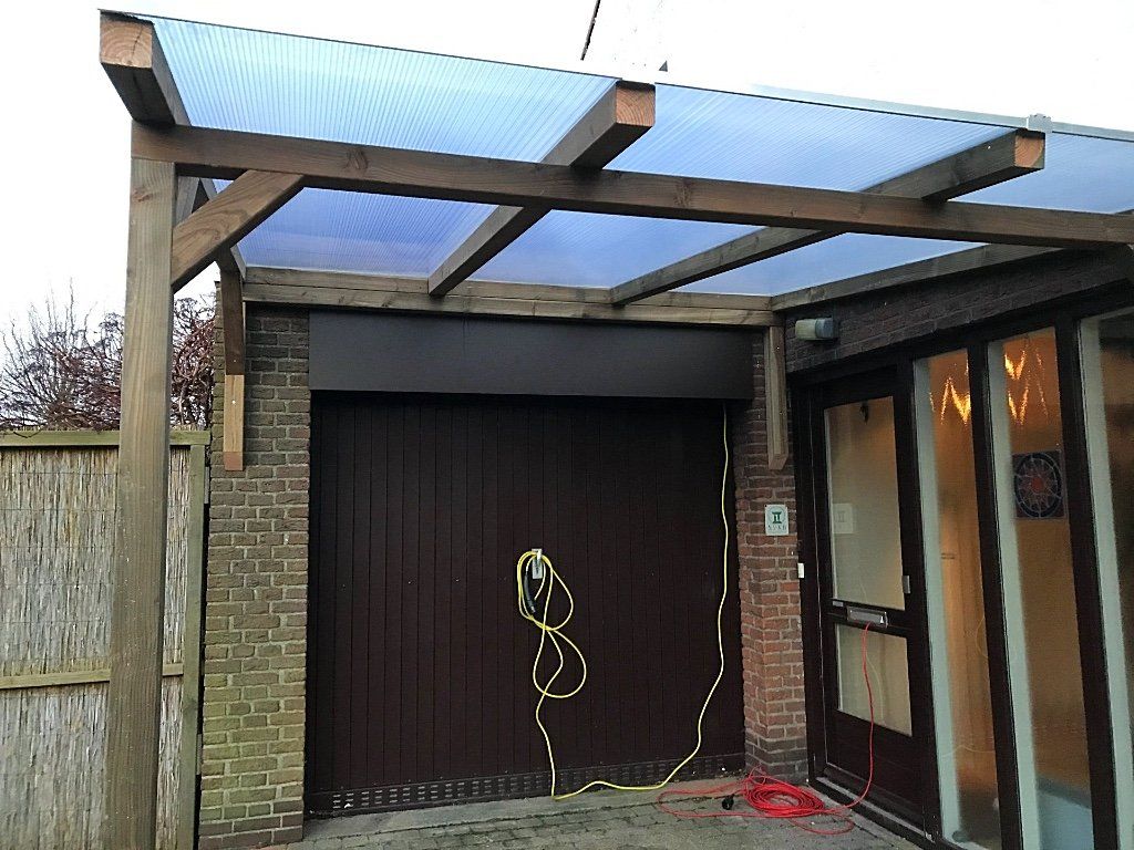 Houten carport boven een bruine garagedeur. Doorzichtig blauw dak, bakstenen muur en een zijdeur met glas.