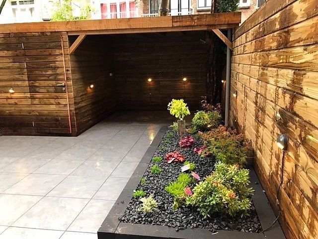 Houten schuurachtige constructie met tuinbed, grijze tegels en verlichting.
