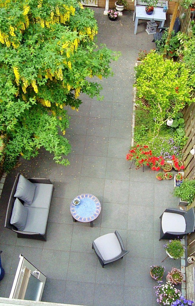 Bovenaanzicht van een gemeubileerd terras met grijze bestrating, een kleine tafel, zitmeubels en een tuin met bomen.