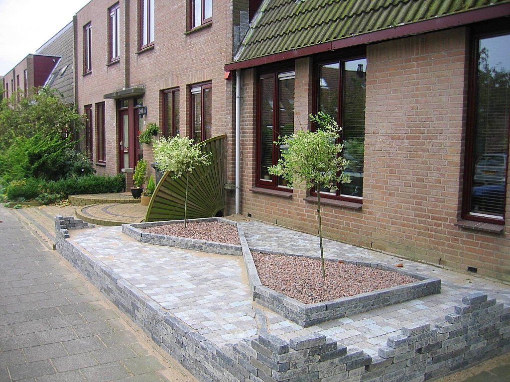 Een lage bakstenen muur met een geometrisch bloembed met twee kleine bomen, naast een bakstenen gebouw.
