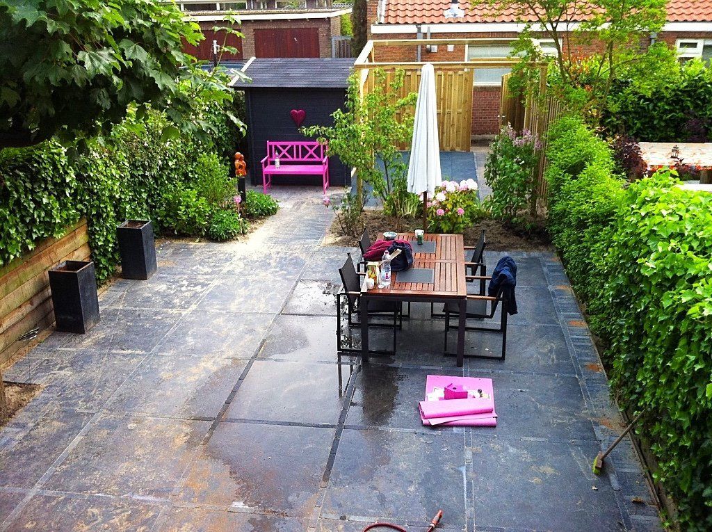 Achtertuin met terras, tafel en stoelen, roze bankje en groene hagen.