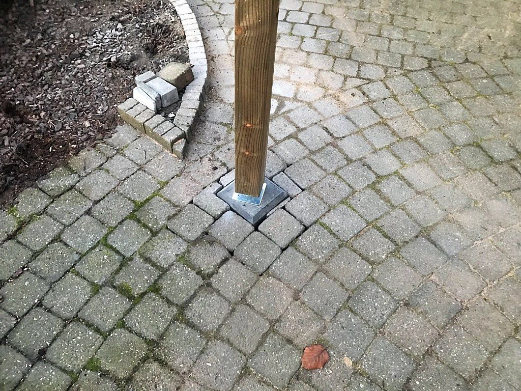 Een houten paal op een vierkante metalen voet, omgeven door grijze kasseien in een buitenomgeving.
