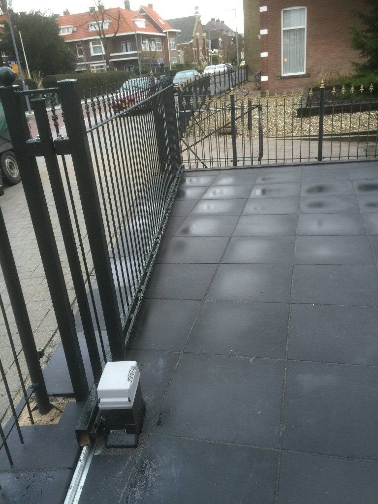 Een zwarte metalen toegangspoort, gedeeltelijk open, met een automatische opener, donker wegdek en huizen op de achtergrond.