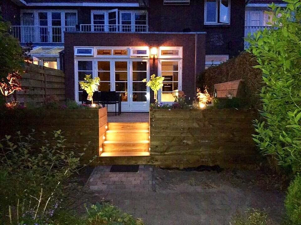 Achtertuin 's nachts, houten trappen die leiden naar een huis met verlichte deuren, omgeven door groen gebladerte en tuinverlichting.