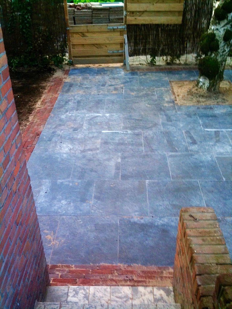 Een stenen terras leidt naar een achtertuinpoort met blauwgrijze stenen bestrating, omzoomd door bakstenen en een houten schutting.