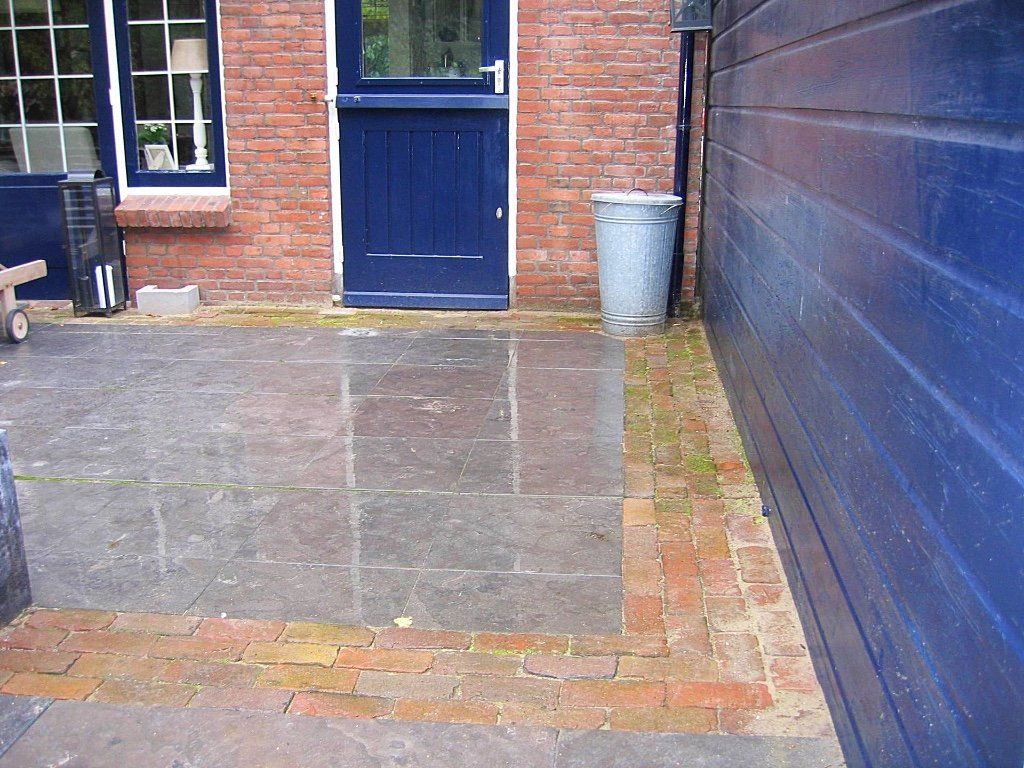 Bakstenen terras met blauwe deur en muur. Rechts van de deur staat een metalen vuilnisbak.