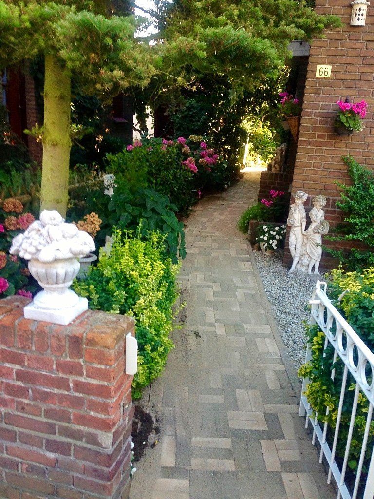 Een bakstenen pad leidt naar de ingang van een huis, omzoomd met groen en bloempotten. Een decoratieve urn staat op een bakstenen pilaar.