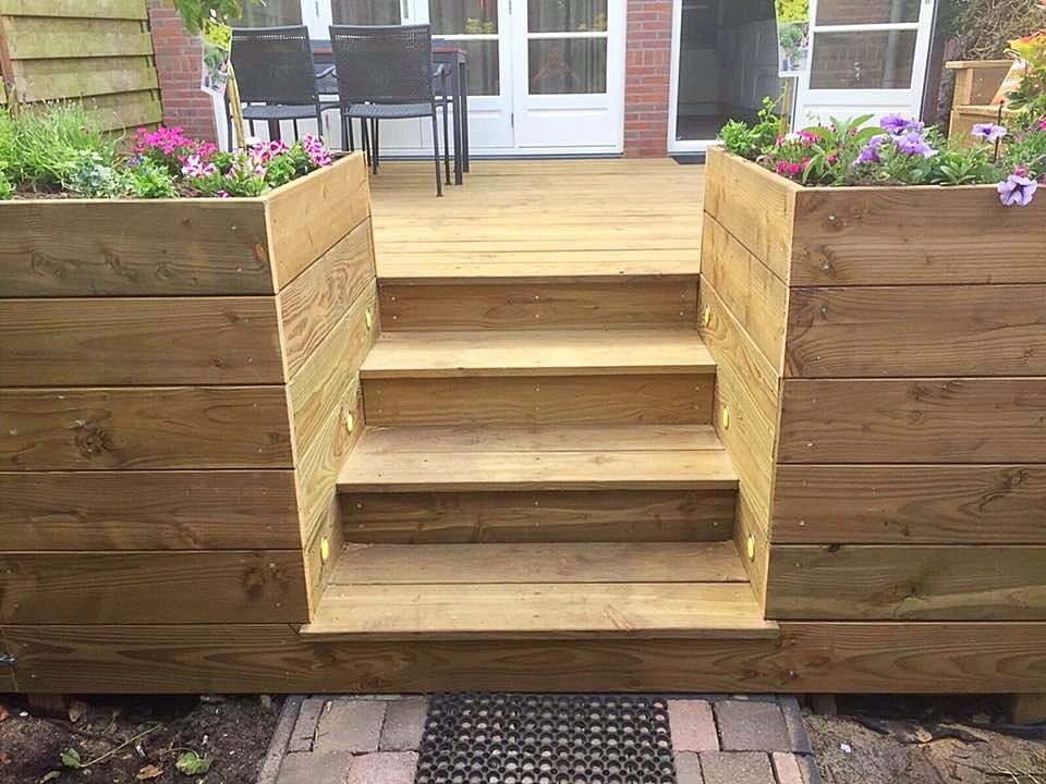 Houten terras met ingebouwde plantenbakken en een trap die leidt naar een patio met een tafel en stoelen.