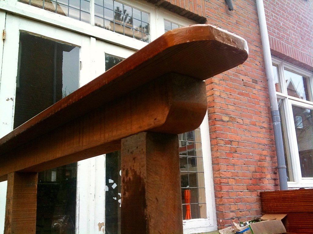 Een houten balustrade, gedeeltelijk bewerkt, voor een bakstenen gebouw met ramen.