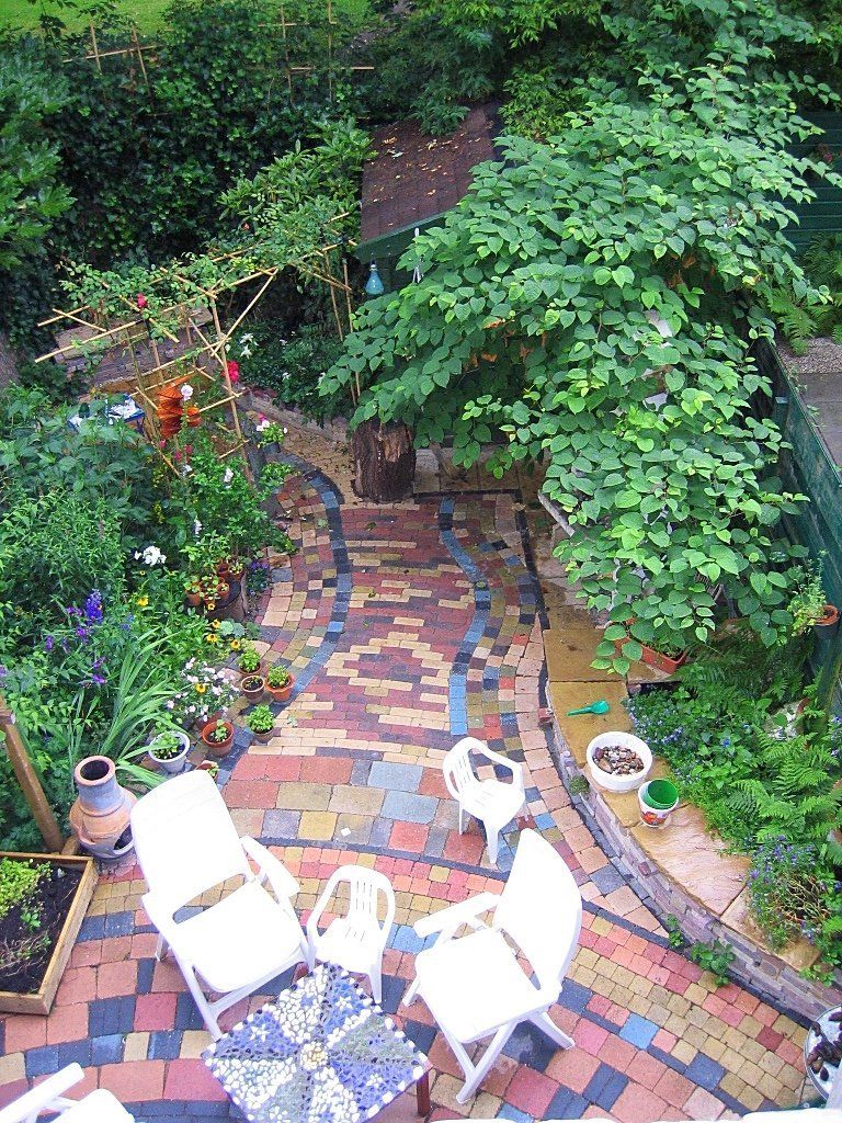 Bovenaanzicht van een kleurrijk bakstenen terras met vier witte stoelen en een klein tafeltje, omgeven door weelderig groen en bloemen.
