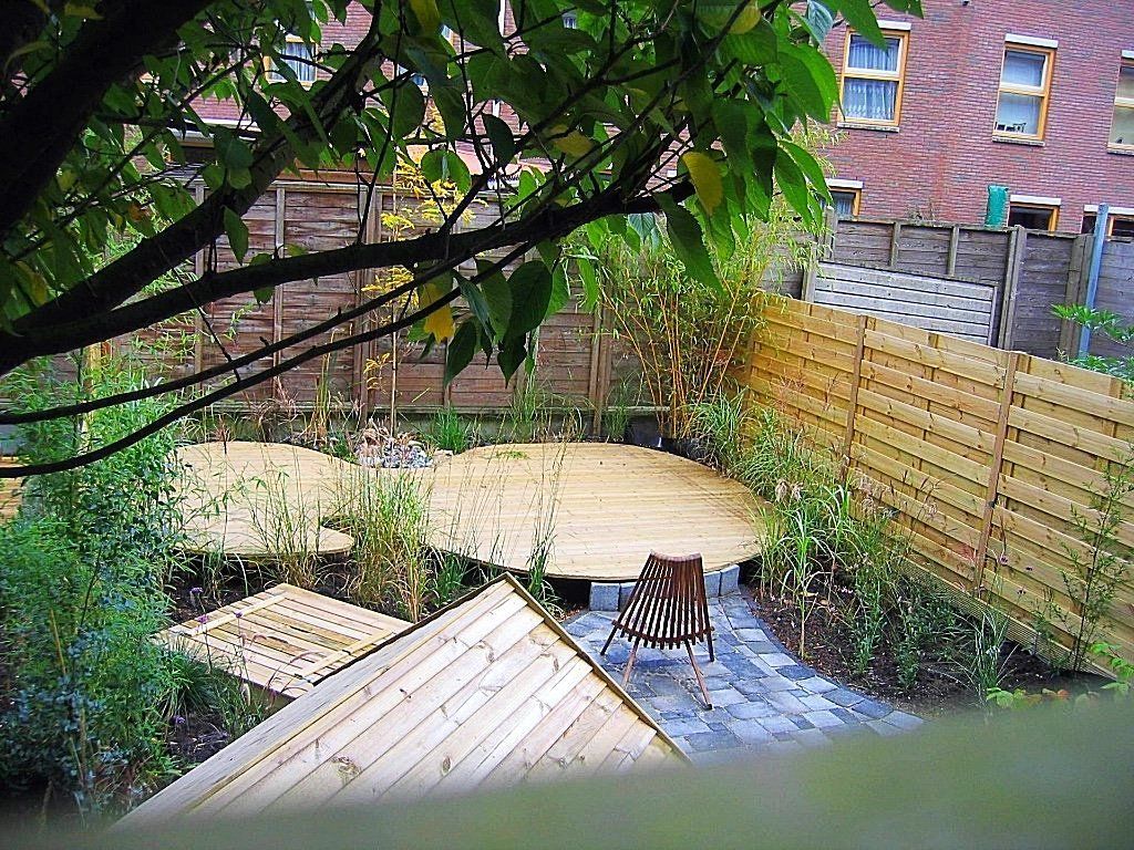 Achtertuin met houten vlonders, bamboe en een bakstenen gebouw op de achtergrond. Een tuinstoel staat op een pad.