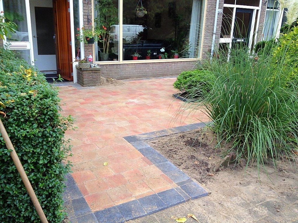 Een stenen terras en een pad leiden naar een huis met ramen en groen, omlijst door struiken en gras.