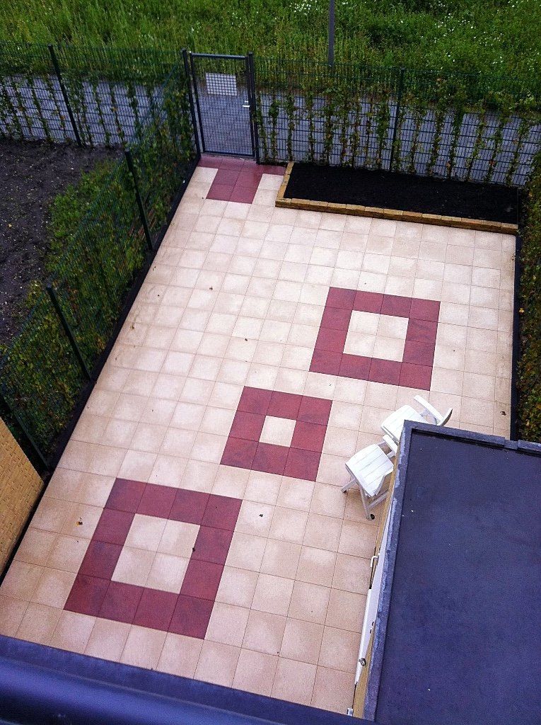 Terras bestraat met beige en kastanjebruine tegels. Rechthoekige ruimte omgeven door een heg en een schutting.