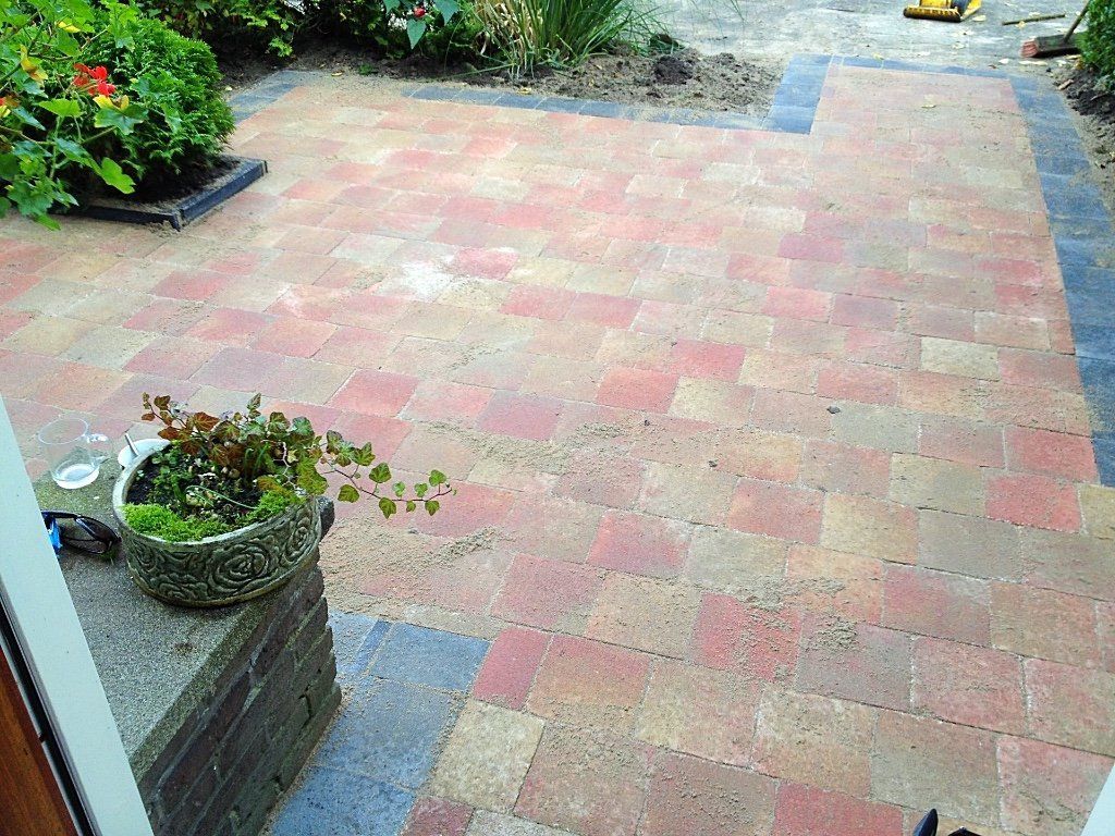 Bakstenen terras met roodbruine bestrating, grenzend aan donkere bestrating, naast een bloembed en een voordeur.