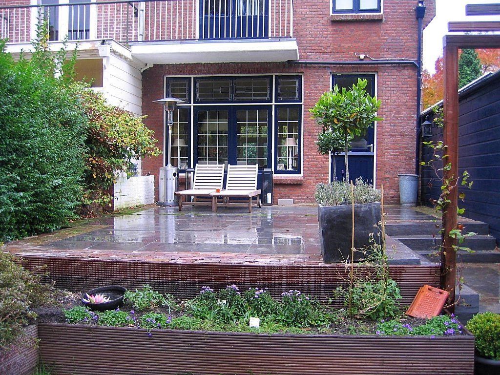 Terras in de achtertuin met een stenen huisje, een houten bank en een bloembed.