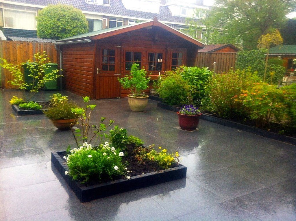Houten schuur in een tuin met bloemperken, omgeven door een donker, nat betegeld terras.