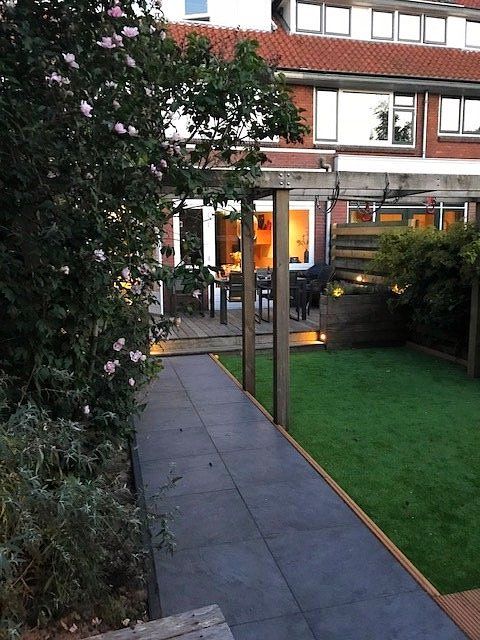 Achtertuin met een donker geplaveid pad, een groen gazon en op de achtergrond een bakstenen gebouw van twee verdiepingen.