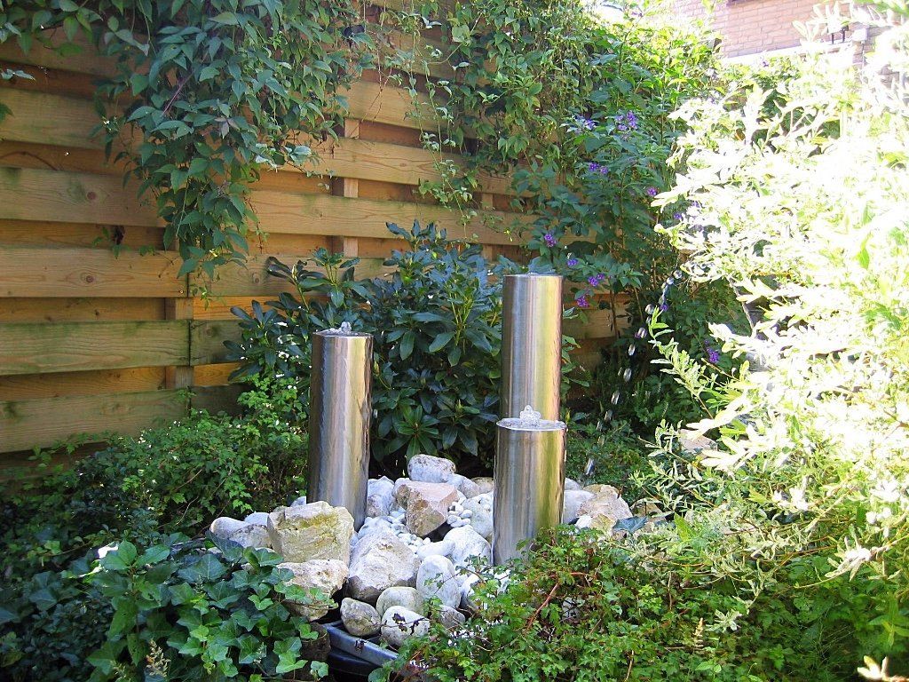 Waterfontein met drie zilveren cilinders, omgeven door rotsen en groen, in een tuinomgeving.