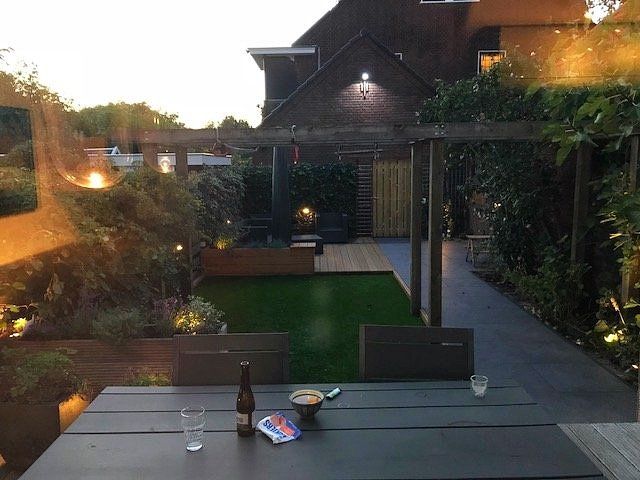 Een terras in de achtertuin met een tafel, drankjes en een klein gazon; verlicht door buitenlampen bij schemering.