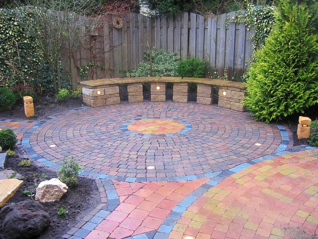 Rond terras met bestrating en een stenen bank, omgeven door beplanting en een schutting.