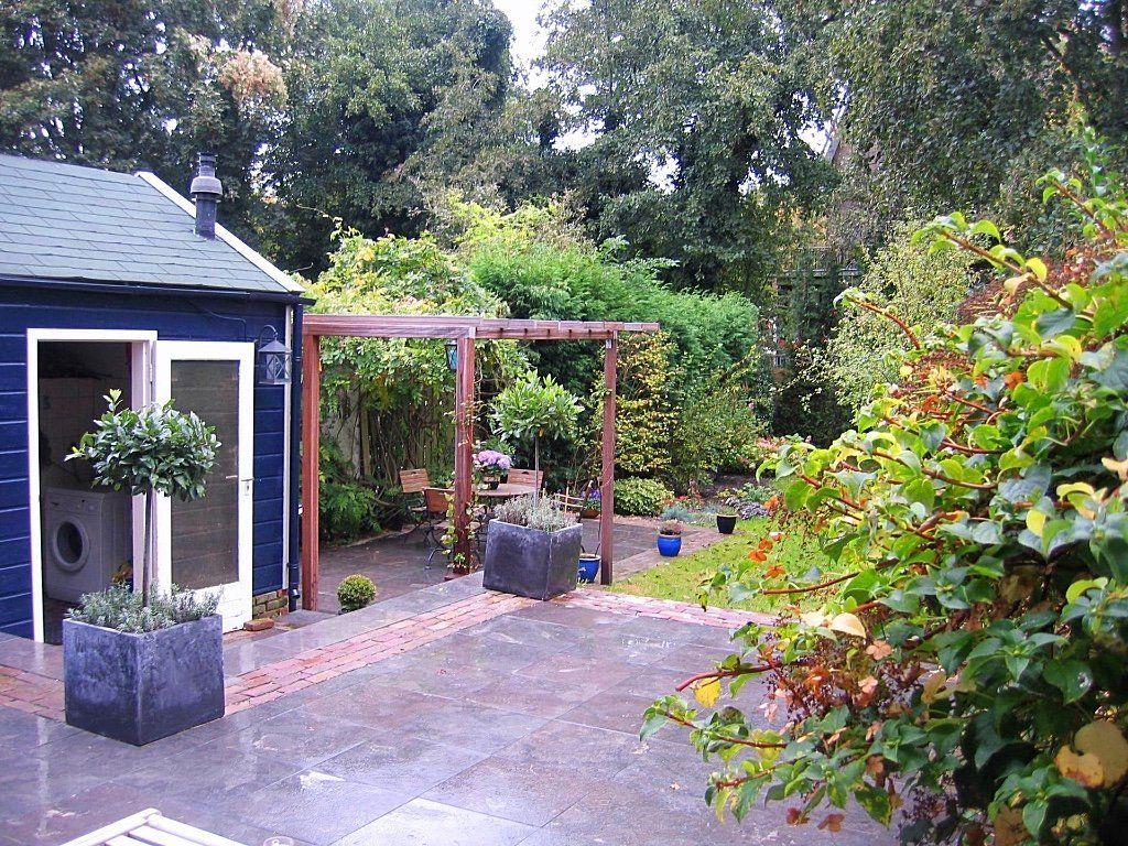 Een terras in de achtertuin met een blauw schuurtje, een pergola en weelderig groen op een bewolkte dag.
