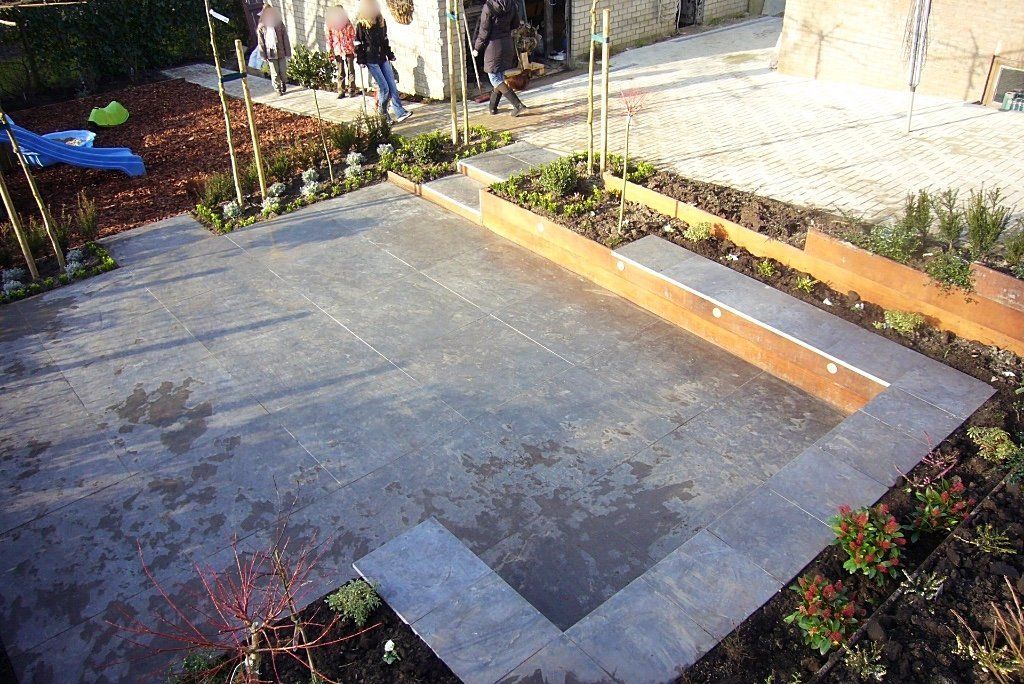 Een moderne achtertuin met een stenen terras, verhoogde bloembedden en trappen. Op de achtergrond zijn enkele mensen te zien.