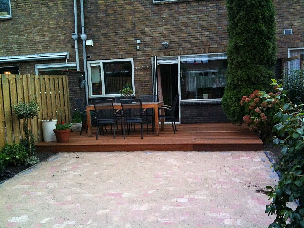Achtertuin met een stenen terras, een houten vlonder en een eettafel; op de achtergrond is een stenen muur met ramen te zien.