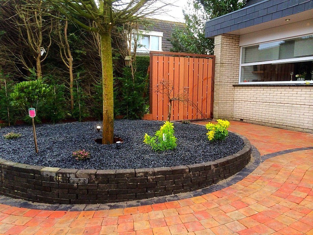 Rond bloembed met een boom en donkere stenen, omzoomd door bakstenen, naast een huis met een bakstenen oprit.