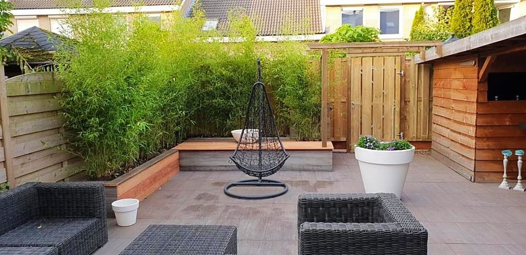 Terras met rieten meubels, een hangstoel, houten schutting en weelderig groen.