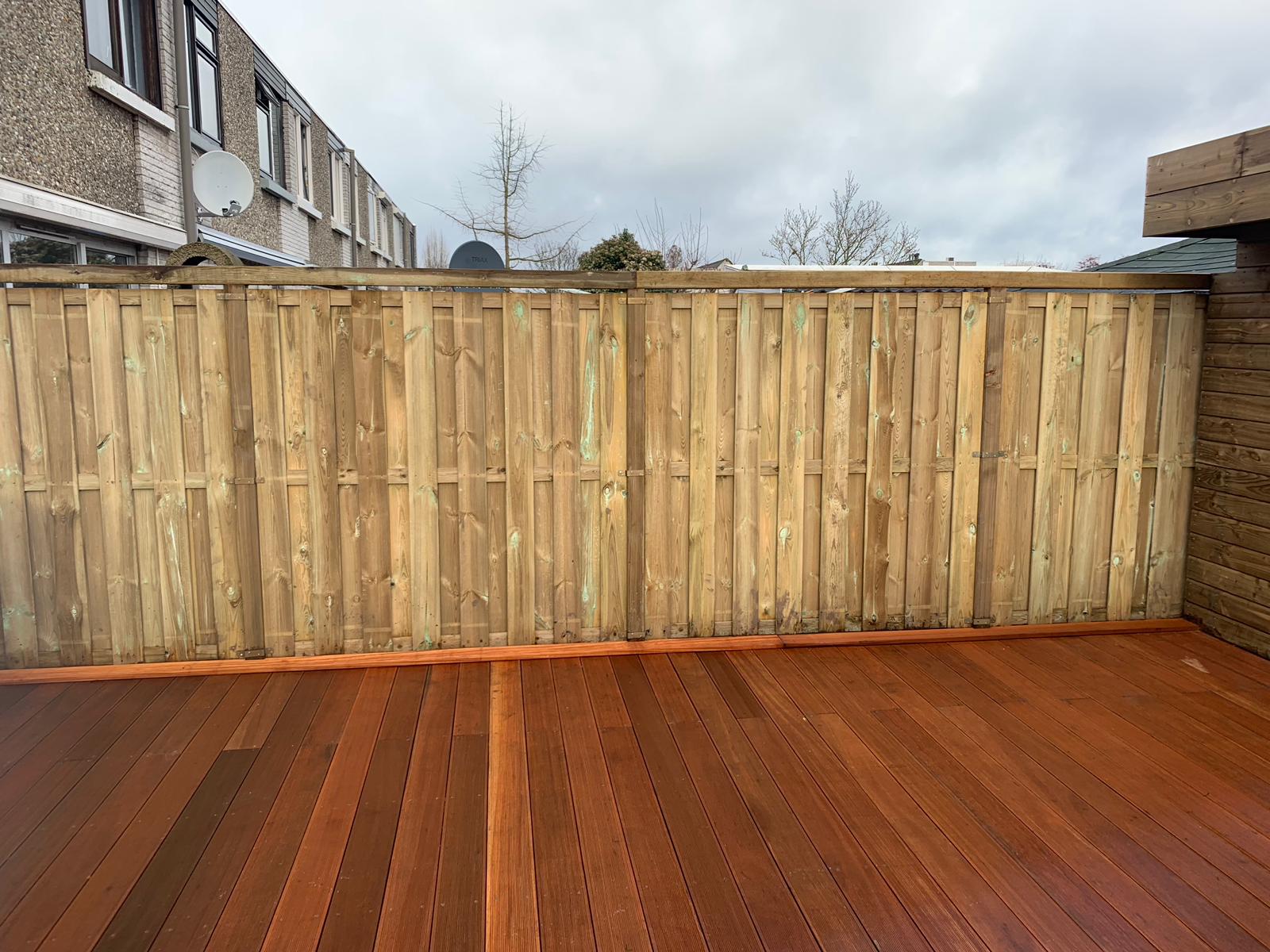 Houten terras en schutting, waarschijnlijk buiten, met op de achtergrond een bakstenen gebouw zichtbaar onder een bewolkte hemel.