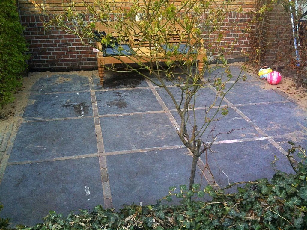 Terras met donkere vierkante tegels, bakstenen muren, een houten bank en een kleine boom.