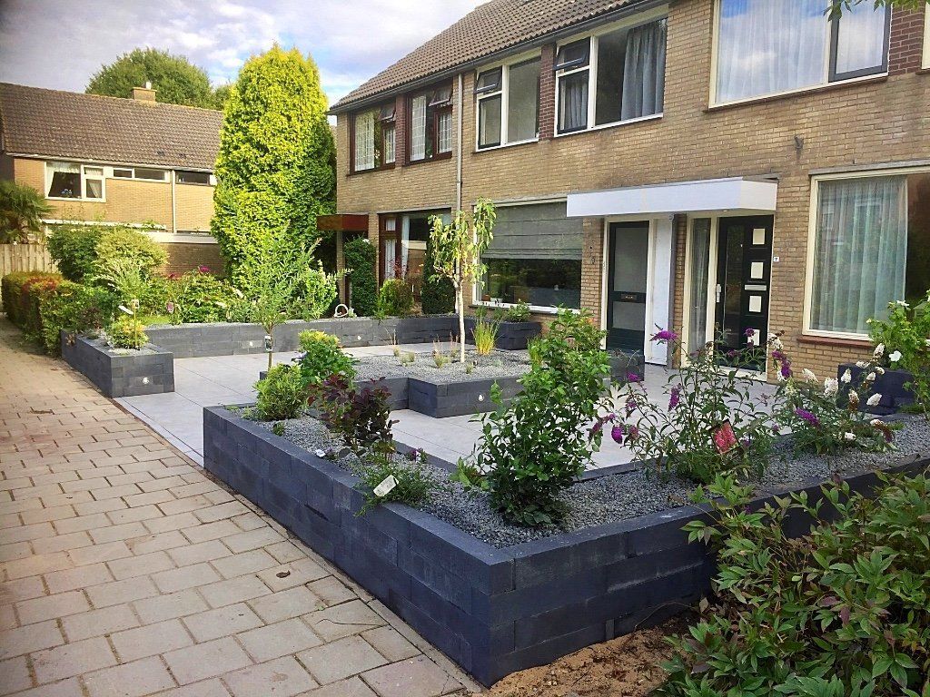 Een moderne, aangelegde voortuin met verhoogde bloembedden, een grijs terras en een twee verdiepingen tellend bakstenen huis.