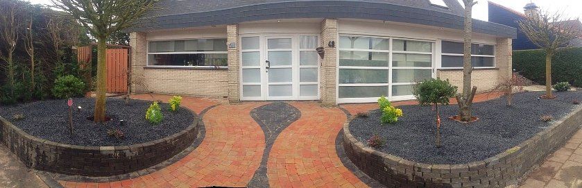 De voorkant van het huis heeft een bakstenen pad dat leidt naar dubbele deuren en ramen, een tuin met zwarte stenen en bomen.