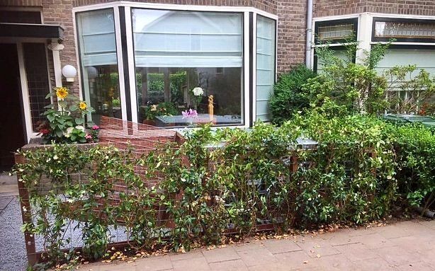 Een bakstenen gebouw met een grote erker, omgeven door een lage heg en groen.