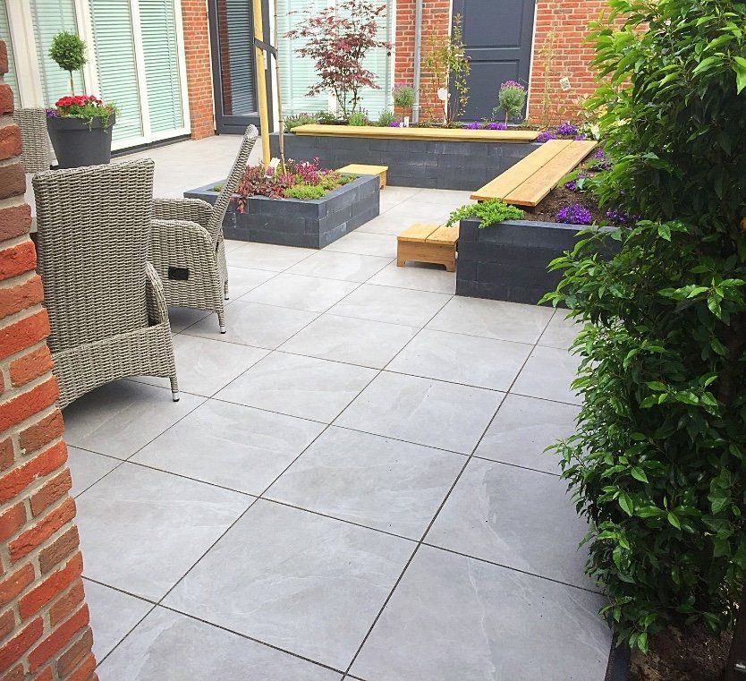 Terras met grijze tegels, rieten meubels en verhoogde plantenbakken met bloemen.