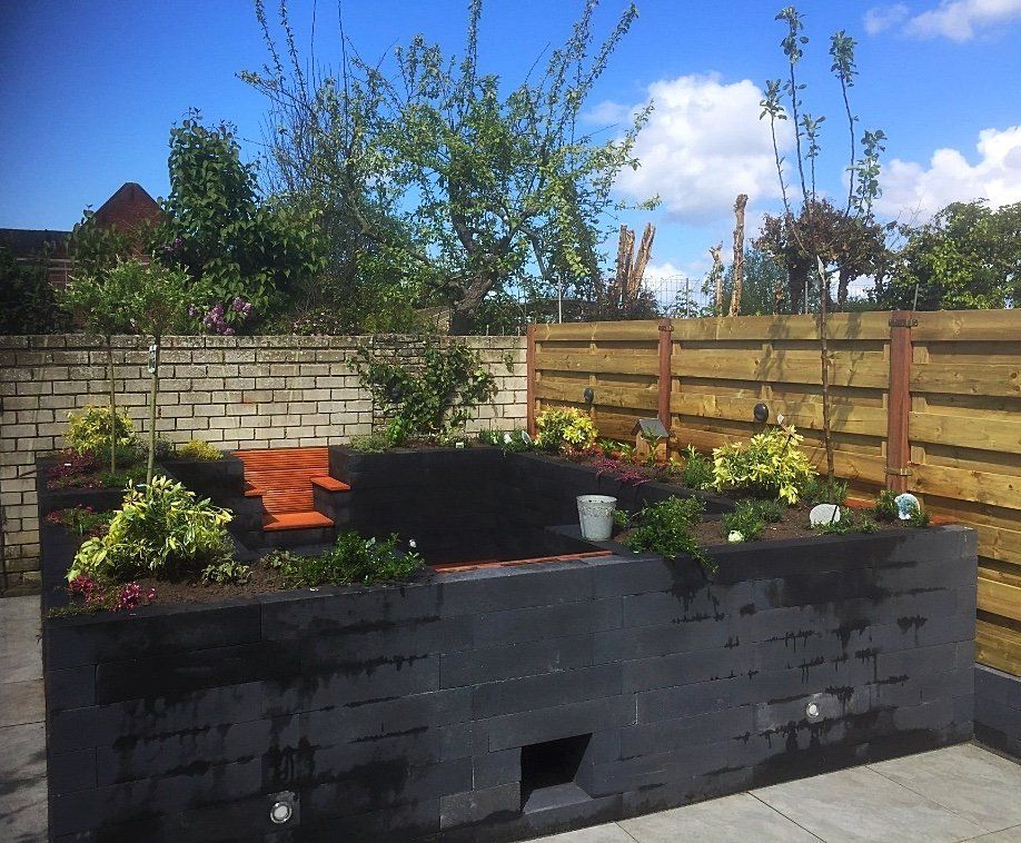 Verhoogd tuinbed met donkergrijze stenen muren, houten schutting, planten en een blauwe lucht.