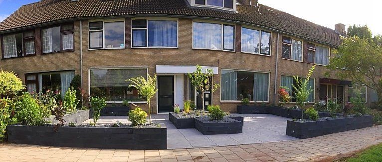 Twee verdiepingen tellend bakstenen gebouw met meerdere ramen, zwarte plantenbakken met groen ervoor. Verhard voetpad.