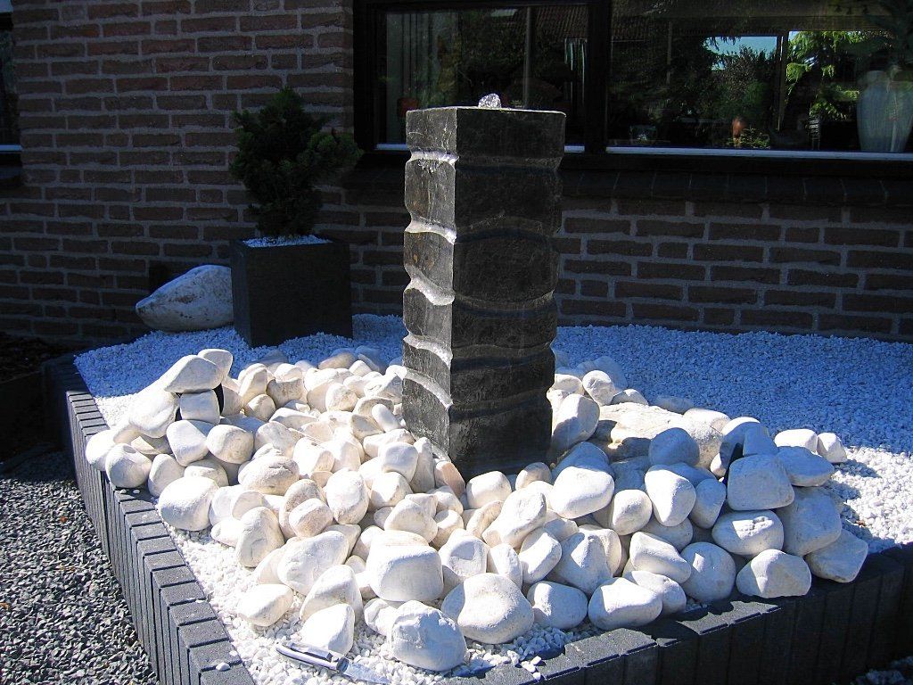 Een waterfontein, omgeven door witte stenen, in een tuin, met een bakstenen muur op de achtergrond.