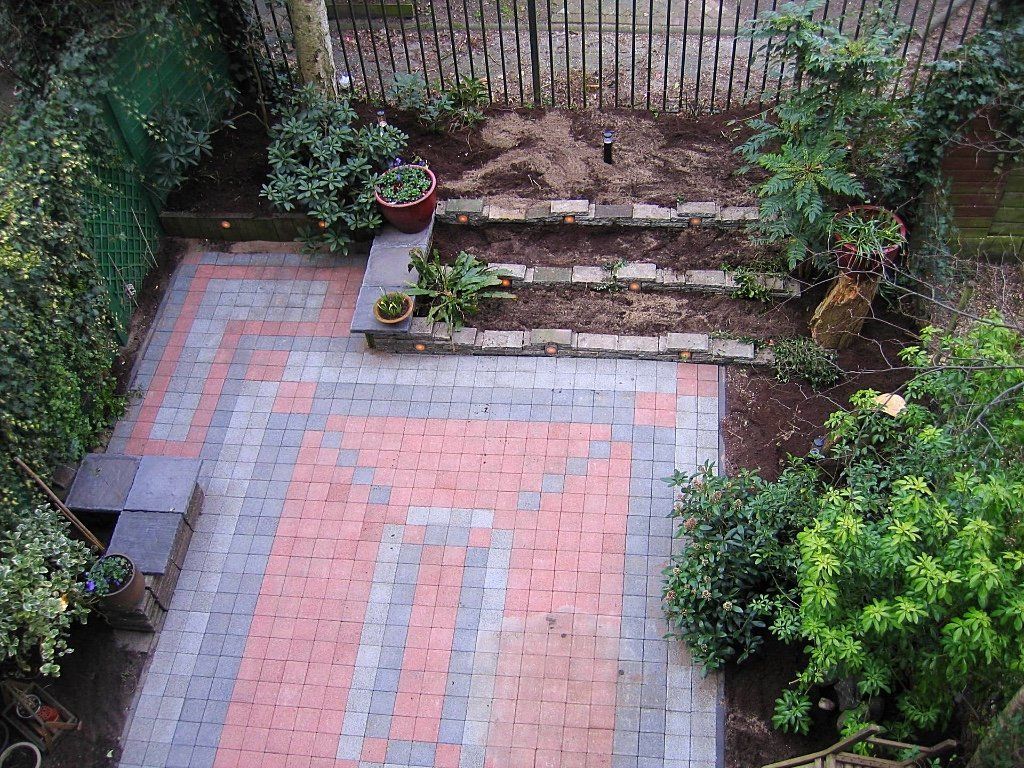 Een terras van bakstenen met een rood en grijs patroon, trappen en een kleine tuin met planten.