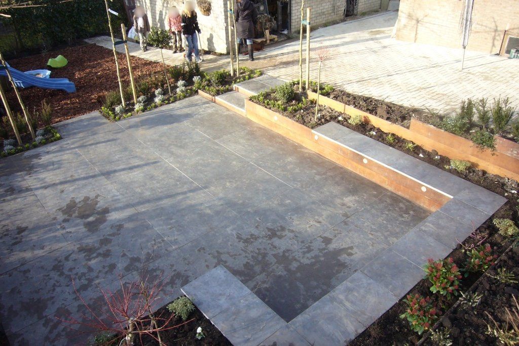 Bovenaanzicht van een moderne achtertuin met stenen terras, houten randen en plantenperken. Op de achtergrond zijn mensen te zien.