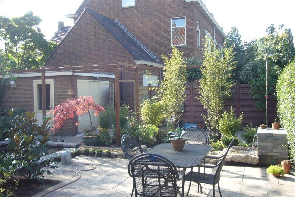 Terras in de achtertuin met een tafel en stoelen, omgeven door bomen, planten en een bakstenen huis.
