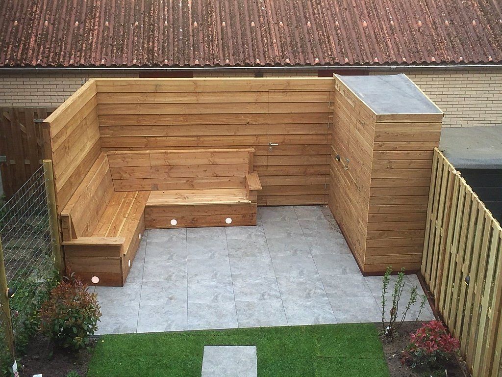 Een houten zithoek in de achtertuin, met grijze tegelvloer, omgeven door houten schuttingen en gras.