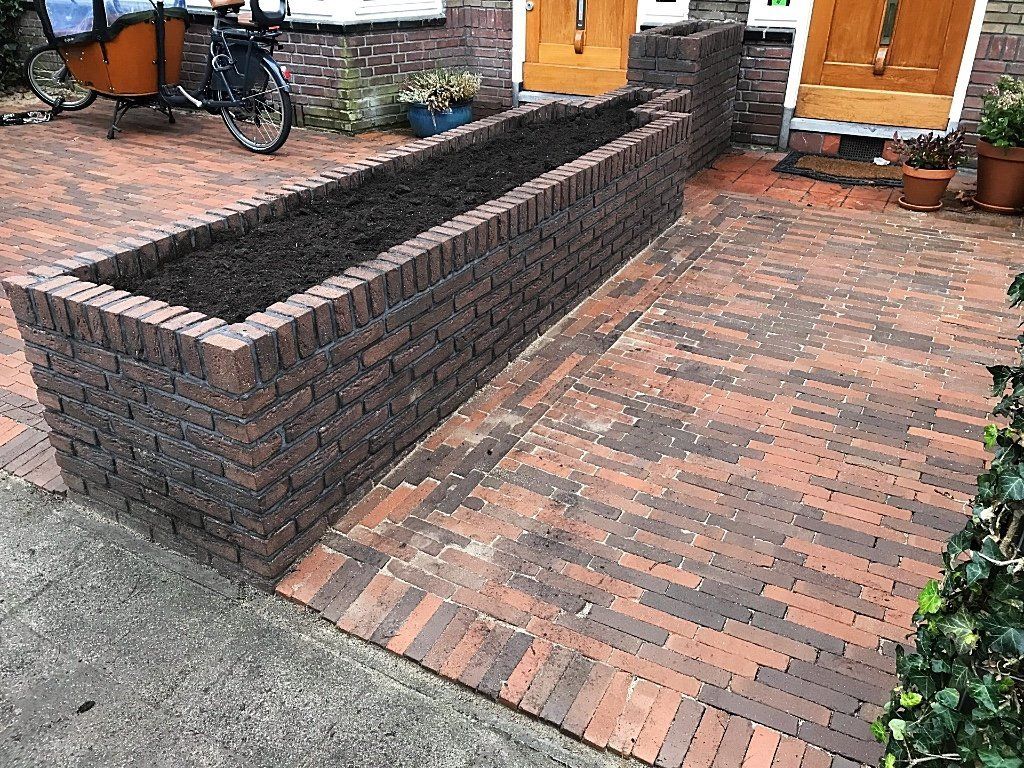 Een bakstenen plantenbak gevuld met donkere aarde staat op een bakstenen terras. Een bakfiets staat er vlakbij geparkeerd.