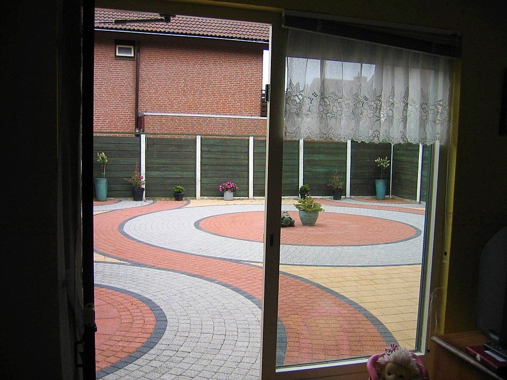 Uitzicht door een glazen schuifdeur op een terras van sierbakstenen met een tuin en een schutting.