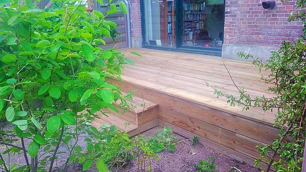 Houten terras met trappen, omgeven door groen, naast een bakstenen gebouw met glazen deuren.