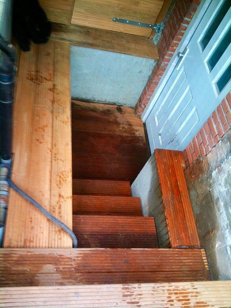 Houten trap die naar beneden leidt naar een deur; leuning aan de linkerkant.