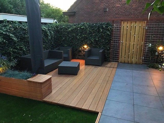 Terras met houten vlonder, grijze tegels, zwart meubilair en een groene haag tegen een bakstenen muur.