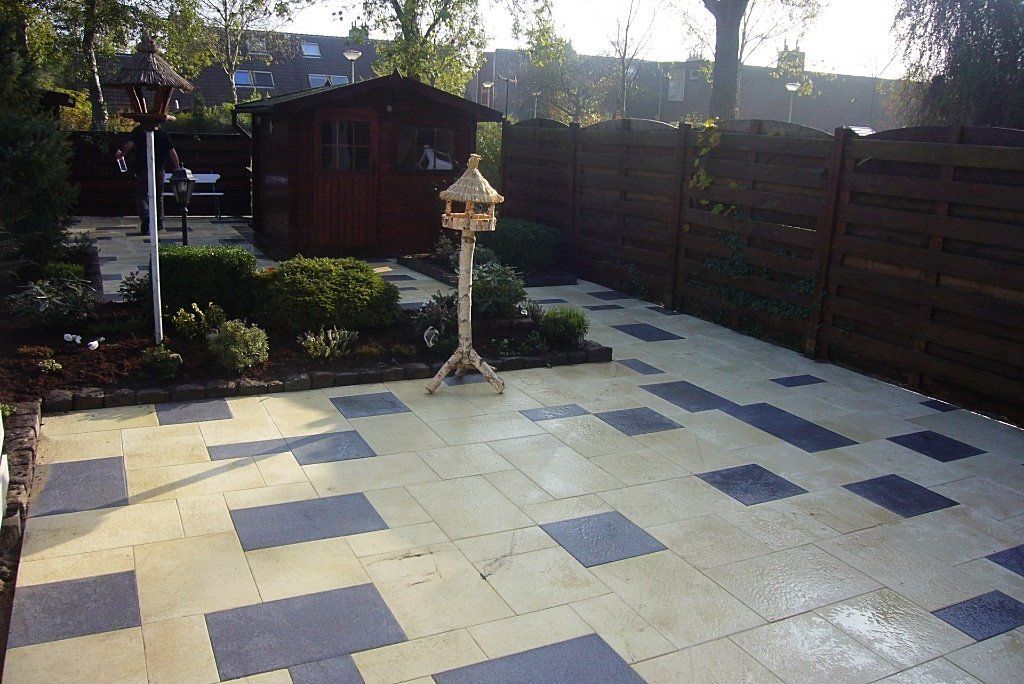 Terras in de achtertuin met lichte en donkere vierkante tegels, houten schutting en schuur, vogelvoederhuisjes.